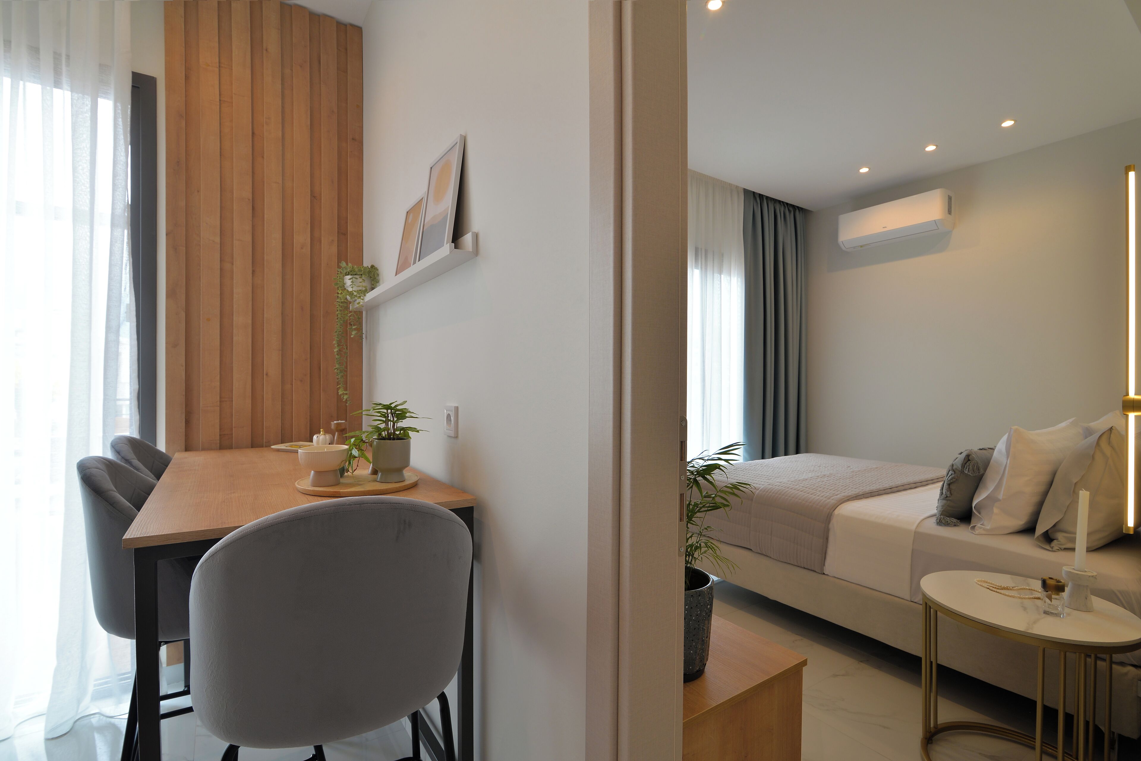Una habitación moderna y bien iluminada con una mesa de comedor de madera, sillas grises, una cama con cabecera blanca y una mesita de noche con lámpara.