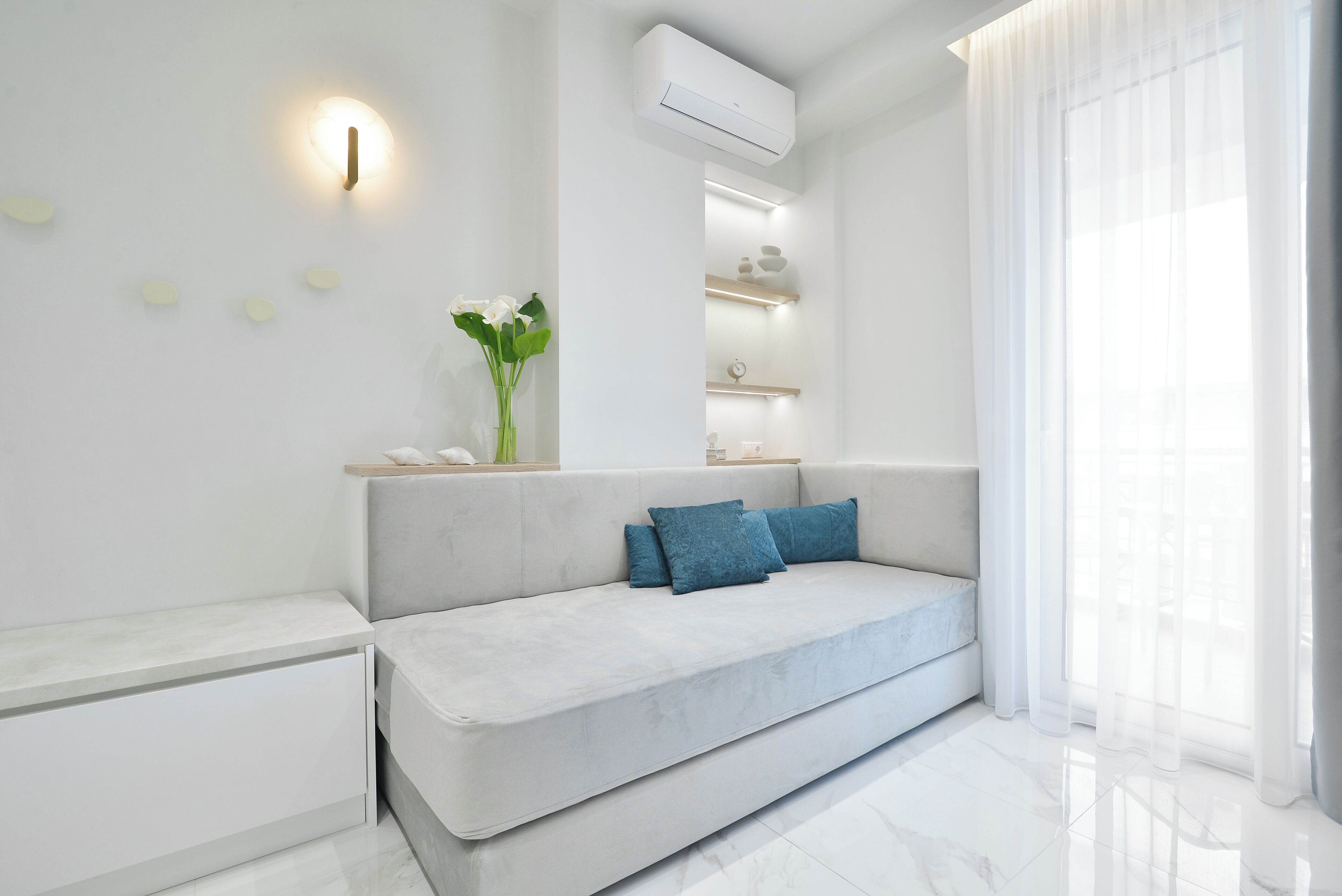 A modern living room with a sofa, a vase with flowers, a wall-mounted light, and a large window with sheer curtains.
