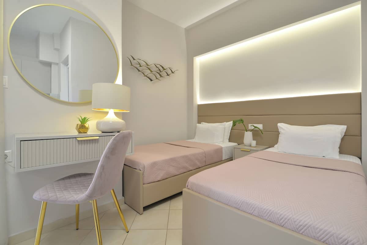 A hotel room with two beds, a desk with a chair, a round mirror, and a bedside table with a lamp.