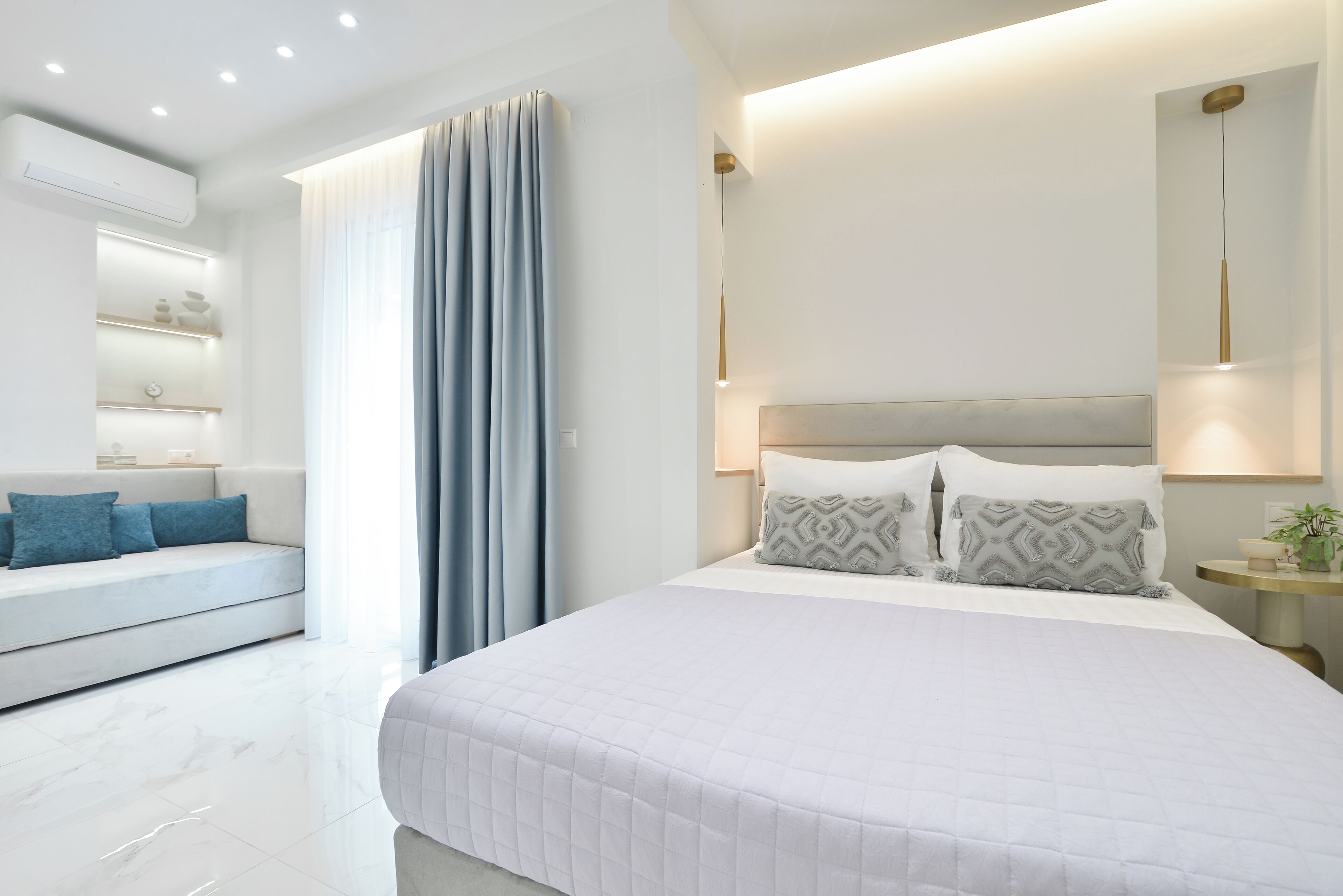 A modern bedroom with a large bed, a sofa, a bedside table with a lamp, and a wall-mounted shelf.