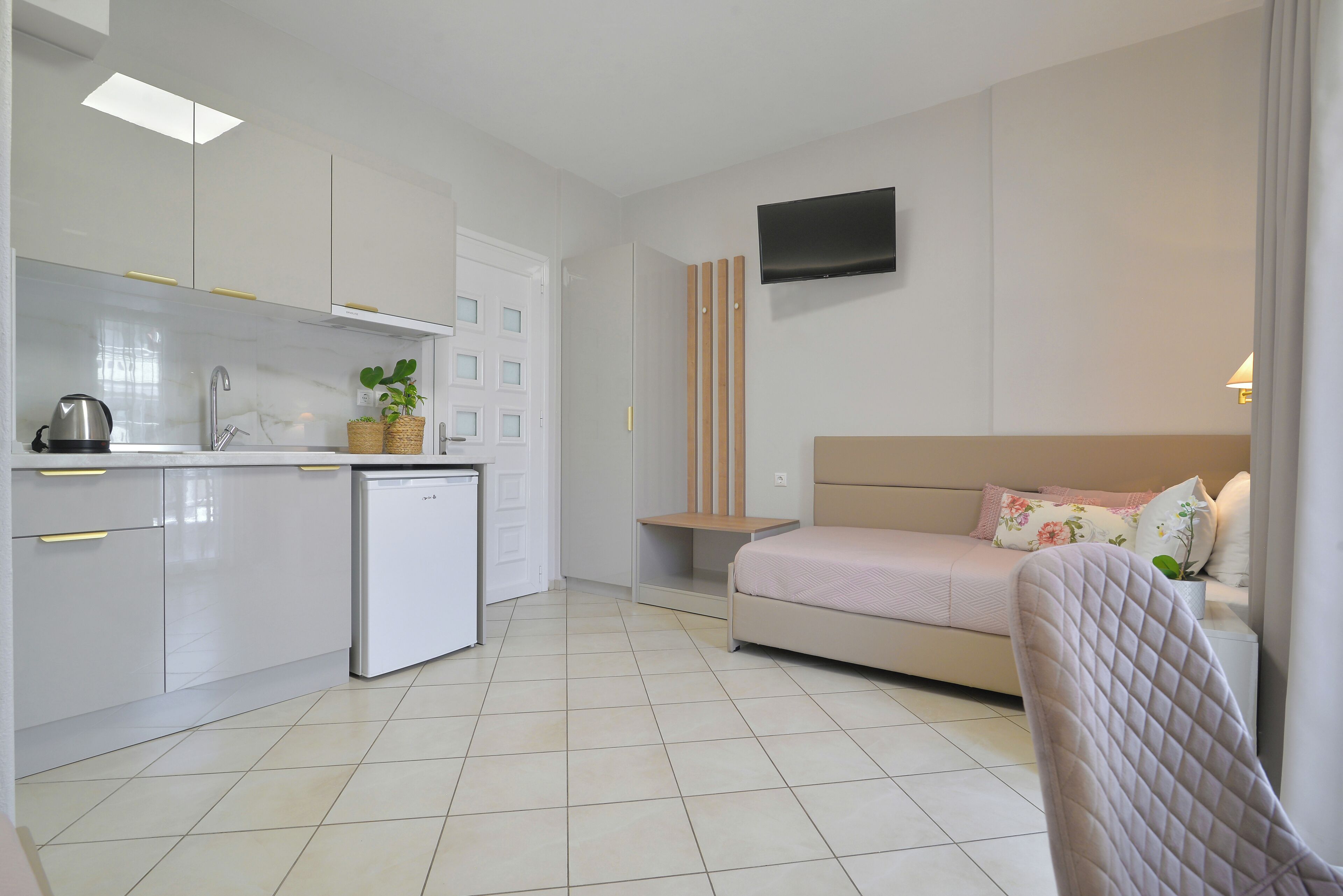 A modern kitchen with white cabinets, a sink, and a dishwasher. There is a small dining table with a plant and a sofa with a TV above it.