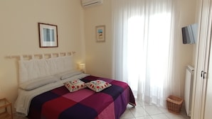 Un dormitorio con cama, lámpara de noche, televisor y ventana con cortinas.