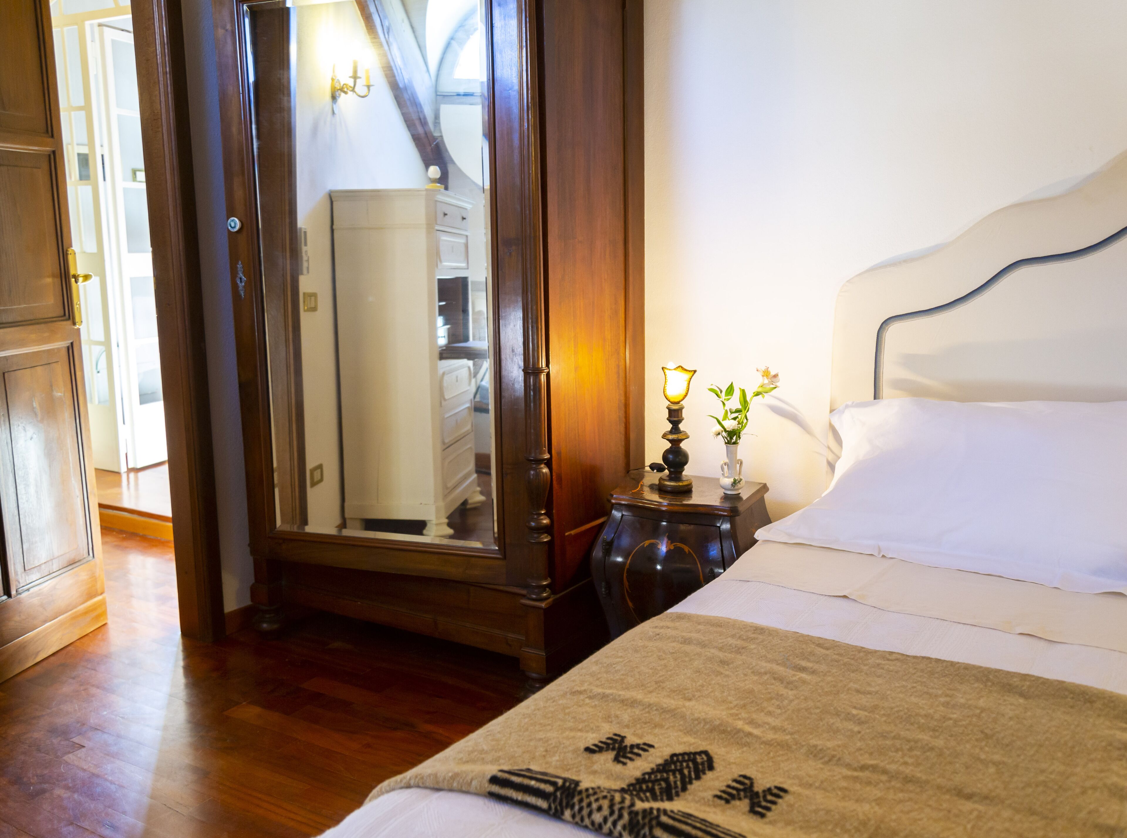 A bedroom with a bed, a nightstand with a lamp, a mirror, and a wooden wardrobe.