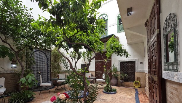 A courtyard with a round table, chairs, potted plants, and a traditional wooden door. - Riad Maizie (Marrakech)