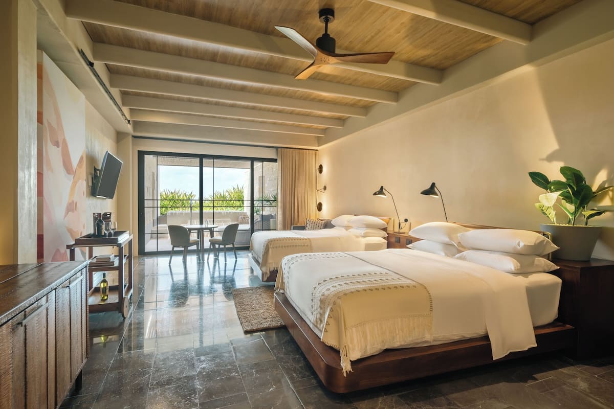 A modern hotel room with two beds, a dining area, a TV, and a large window with a view of greenery.