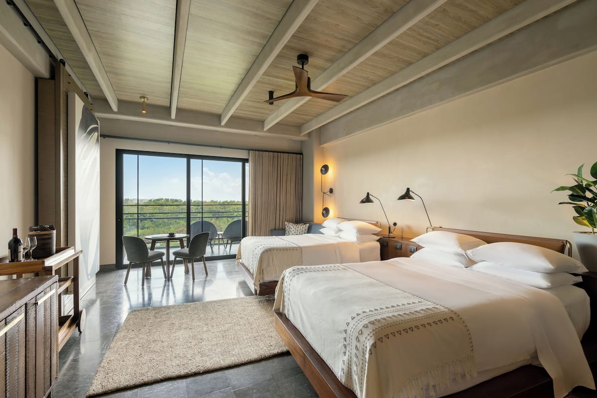 A modern hotel room with two beds, a dining area, and a large window offering a view of the outdoors.