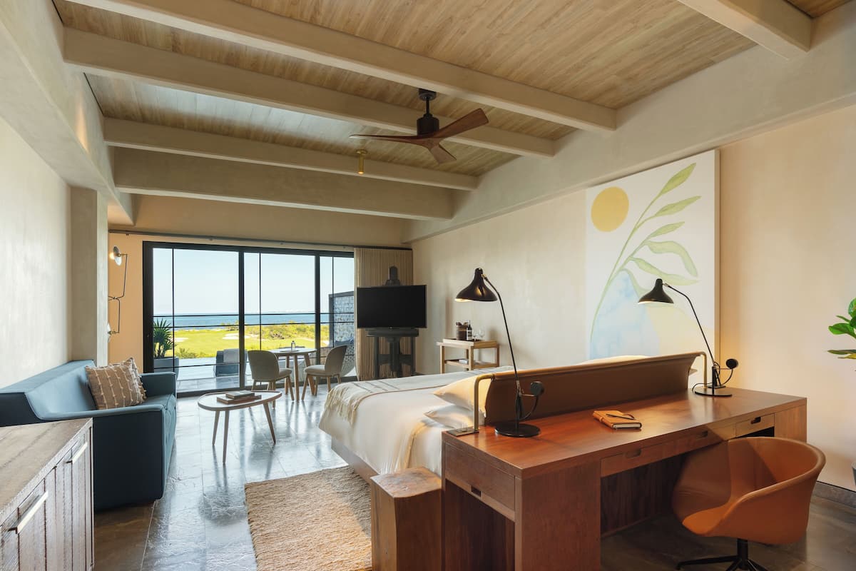 A modern hotel room with a large bed, a desk, a chair, a TV, and a view of the ocean.