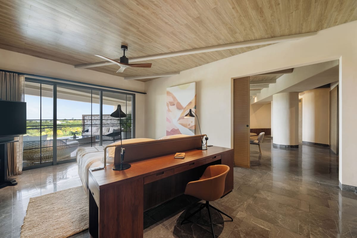 A modern hotel room with a wooden desk, a comfortable chair, a large window with a view, and a ceiling fan.