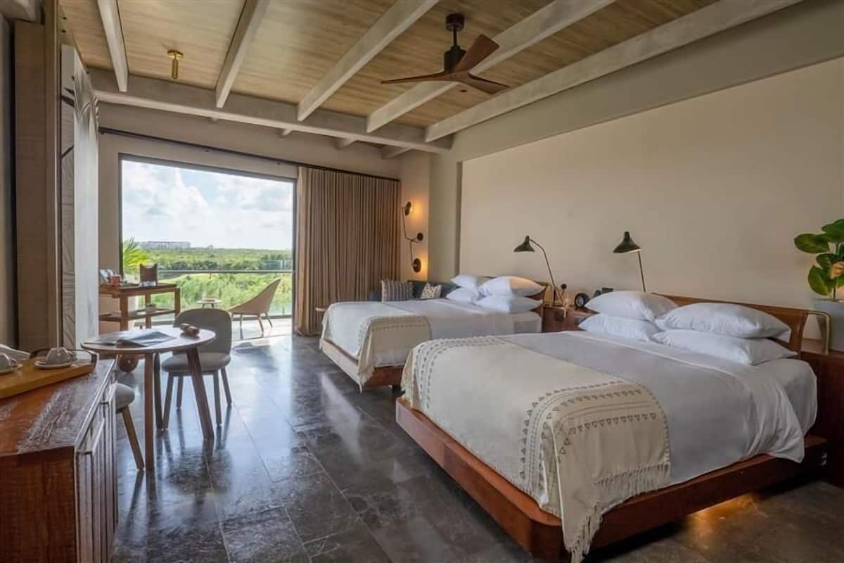 A bedroom with two beds, a desk, and a chair. There is a large window with a view of greenery.