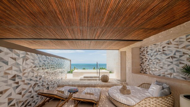 Un espace de détente extérieur, baigné de lumière, avec vue sur la mer, agrémenté d’un plafond en bois, de carreaux muraux aux motifs géométriques et d’un canapé en osier avec des coussins.