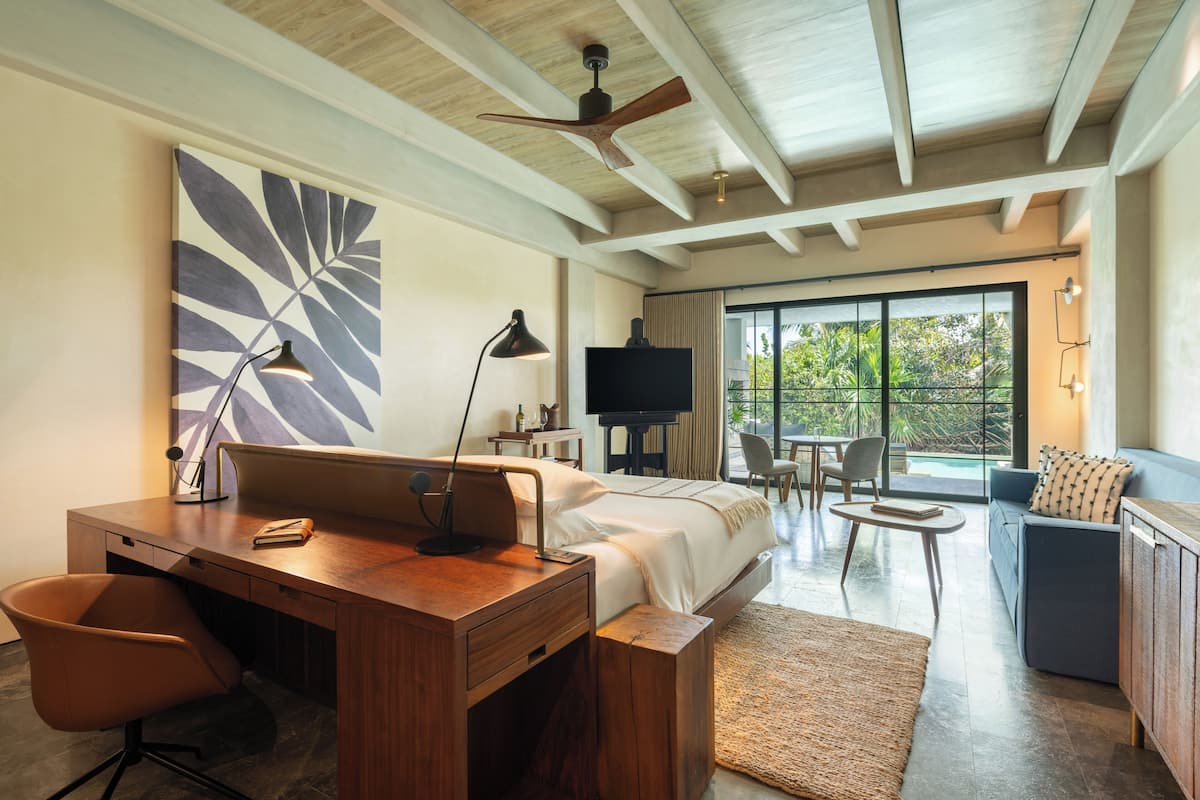A modern hotel room with a wooden desk, a bed, a TV, and a view of the outdoors.