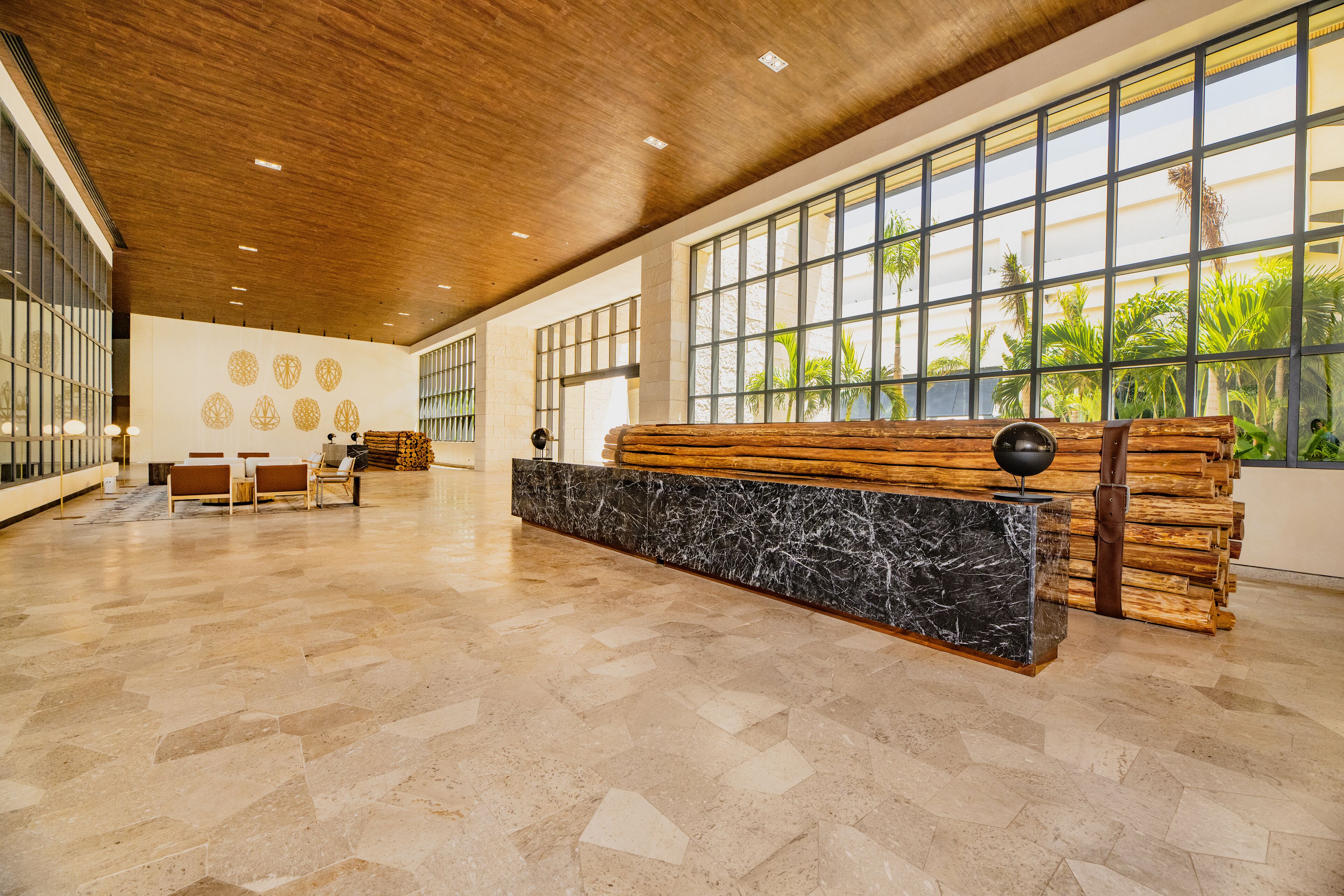 A spacious lobby with a wooden reception desk, marble base, and large windows offering views of palm trees.