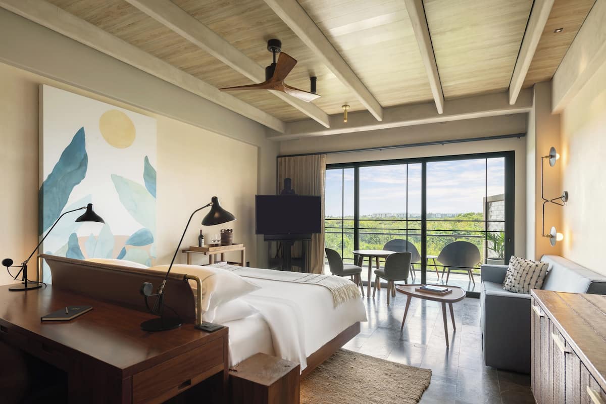 A modern bedroom with a large bed, a desk, a TV, and a balcony with a view of the countryside.