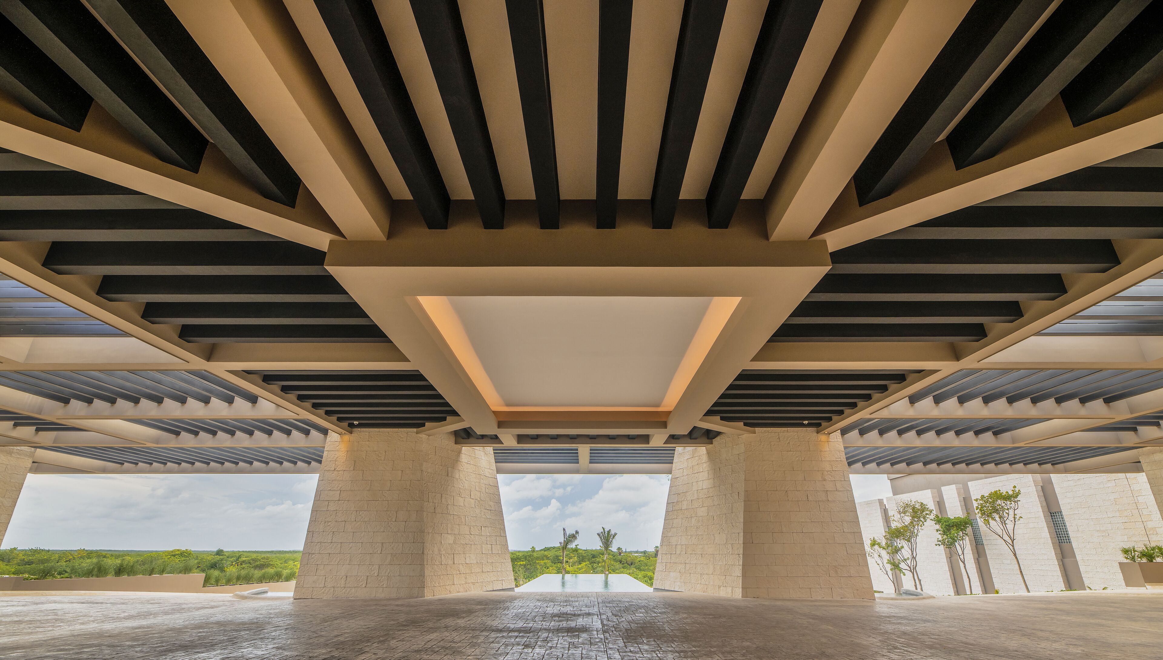 A modern architectural structure with a geometric ceiling design, large pillars, and a view of greenery outside.