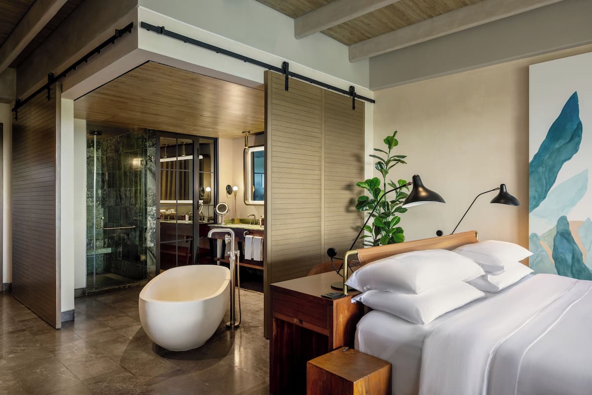 A modern hotel room with a large bathtub, a wooden bed frame, a bedside table, and a large abstract painting on the wall.