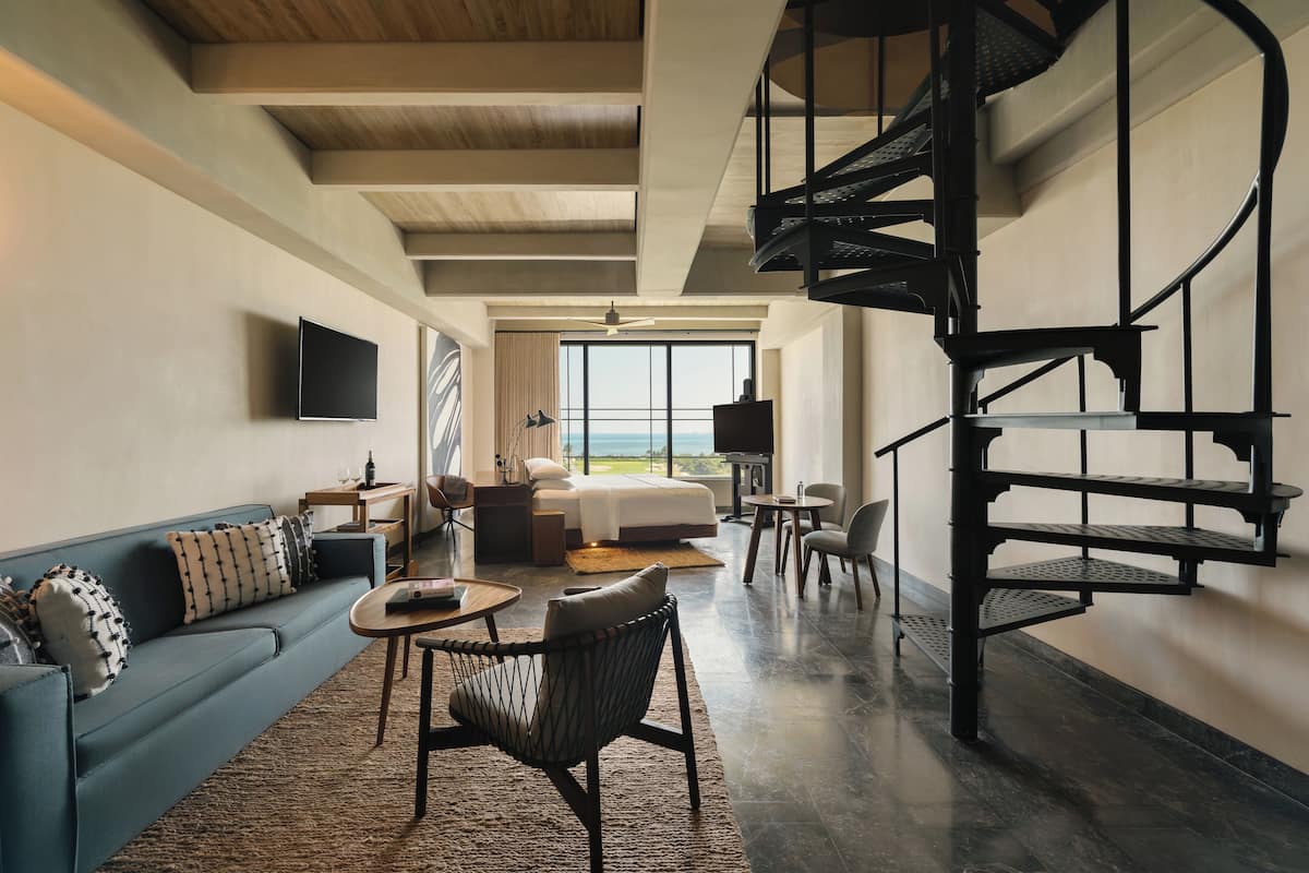 A modern hotel room with a large bed, a sofa, a chair, a small table, a TV, and a spiral staircase.