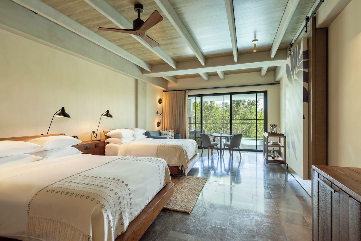 A spacious bedroom with two beds, a dining area, and a large window offering a view of greenery.