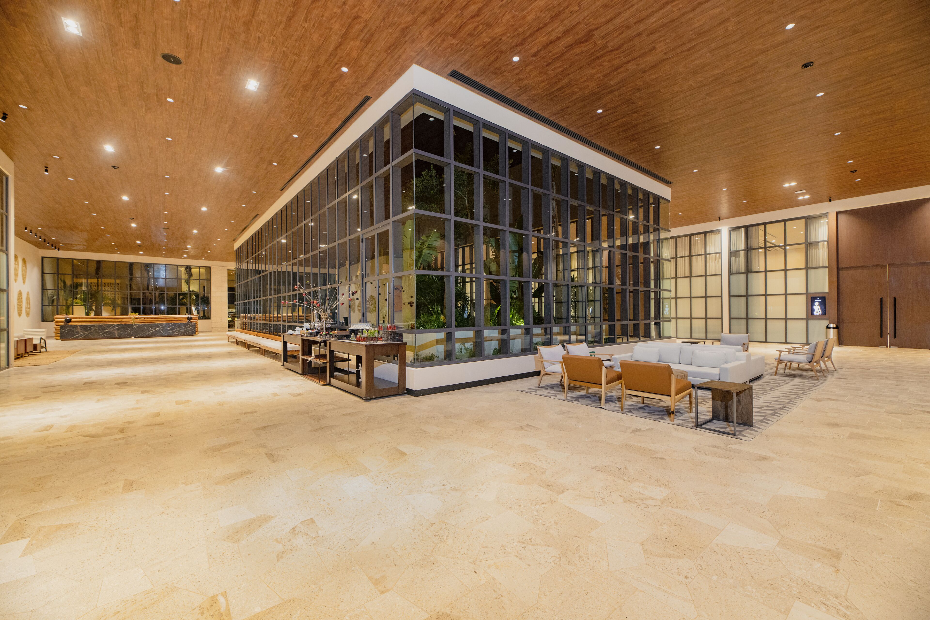 A spacious lobby with a modern design, featuring a large seating area with sofas and chairs, a reception desk, and a large glass wall offering views of indoor plants.