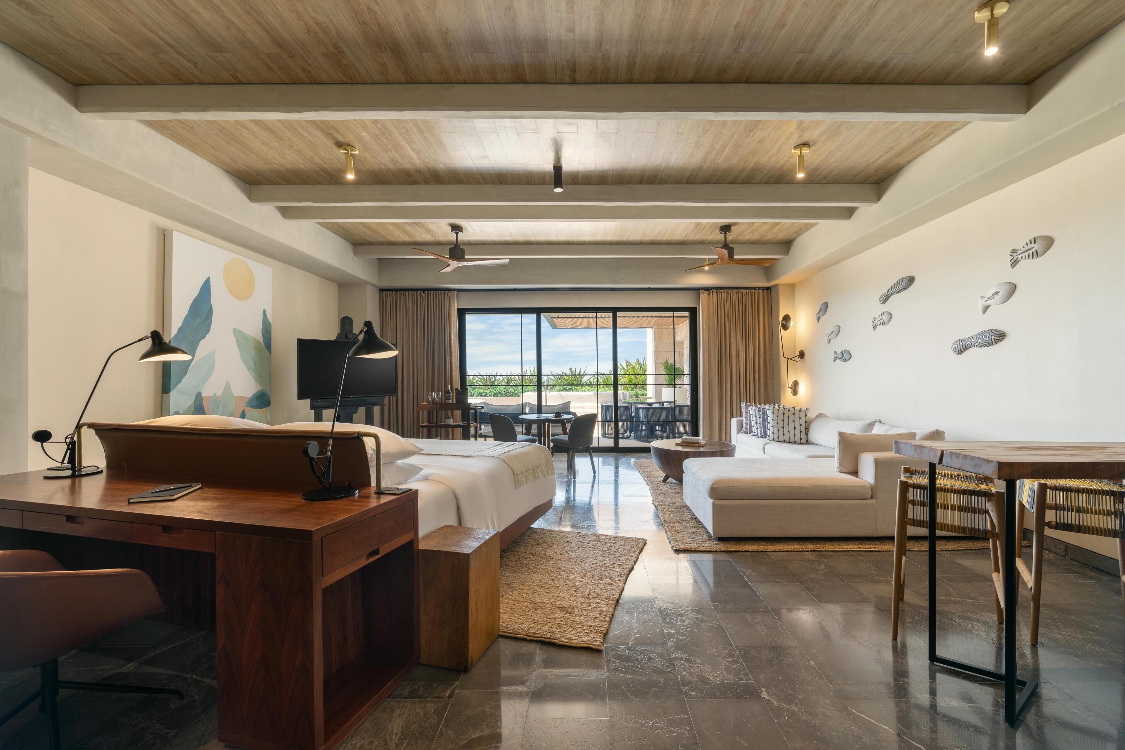 Habitación de hotel moderna con techo de madera, una cama grande, un escritorio con una silla, un sofá, una mesa de comedor con sillas y vistas al exterior a través de amplias ventanas.