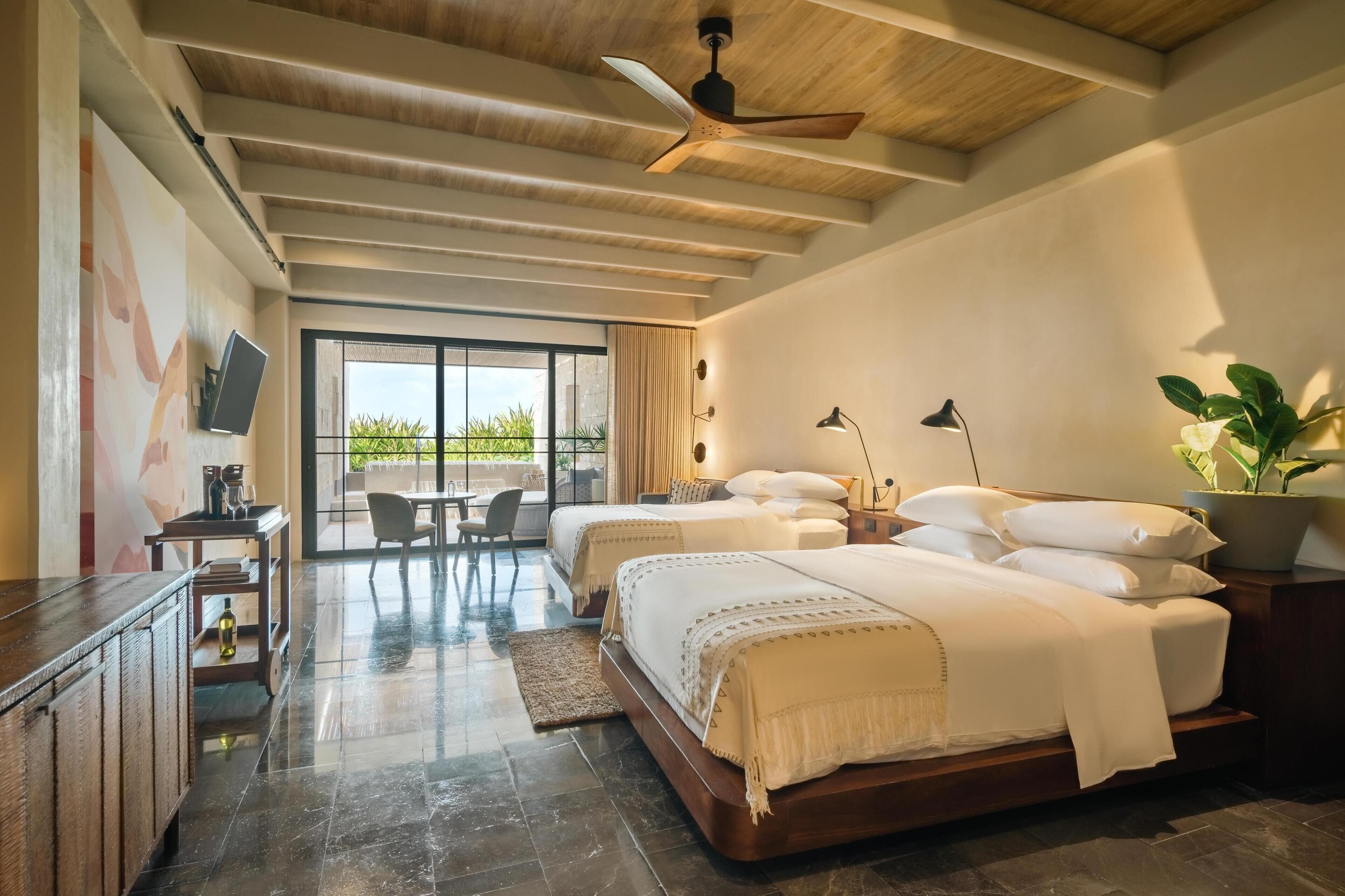 A hotel room with two beds, a desk, a chair, a TV, and a large window with a view of greenery.