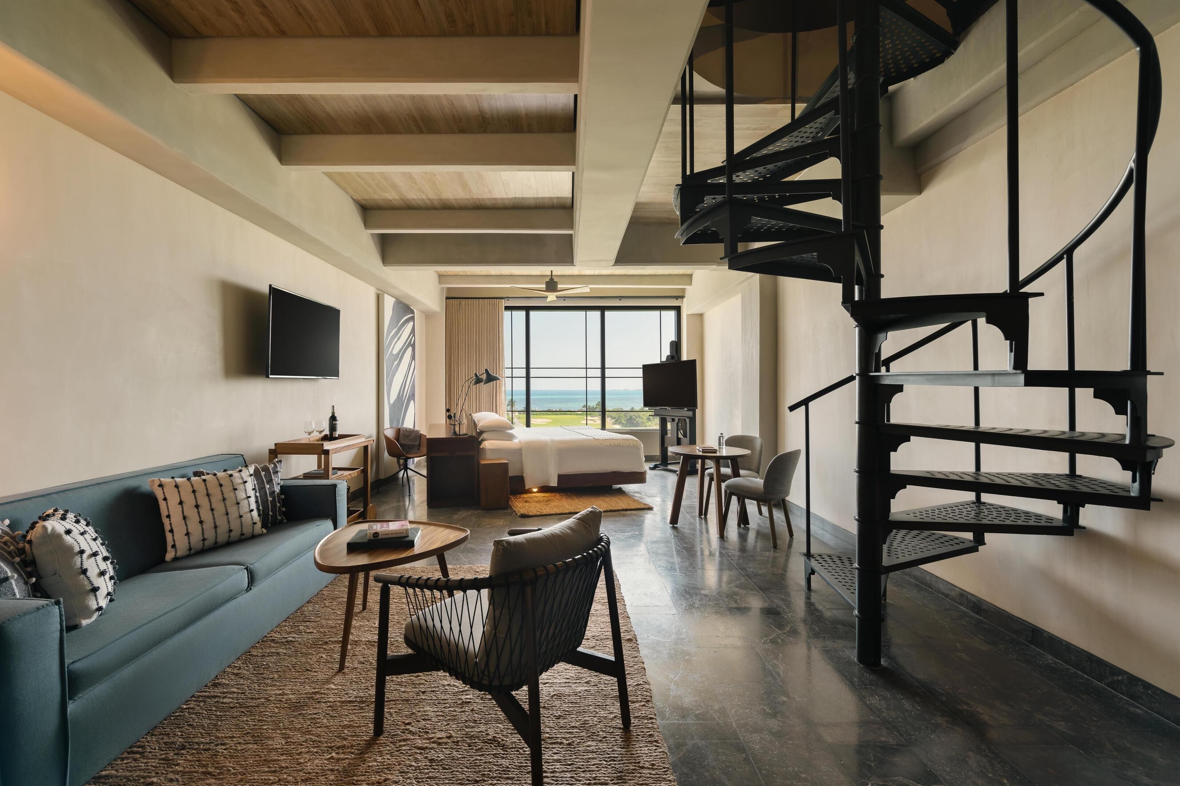 A modern hotel room with a large bed, a sofa, a chair, a desk, and a spiral staircase.
