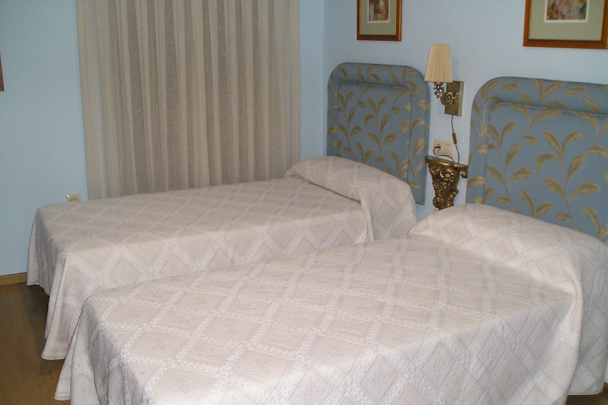 Una habitación con dos camas, una mesita de noche con lámpara y una ventana con cortinas.
