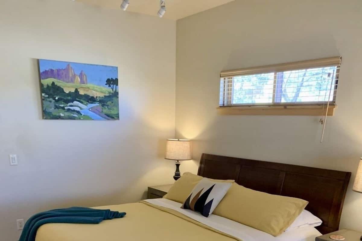 A neatly made bed with a beige bedspread and pillows, a nightstand with a lamp, a painting on the wall, and a window with blinds.