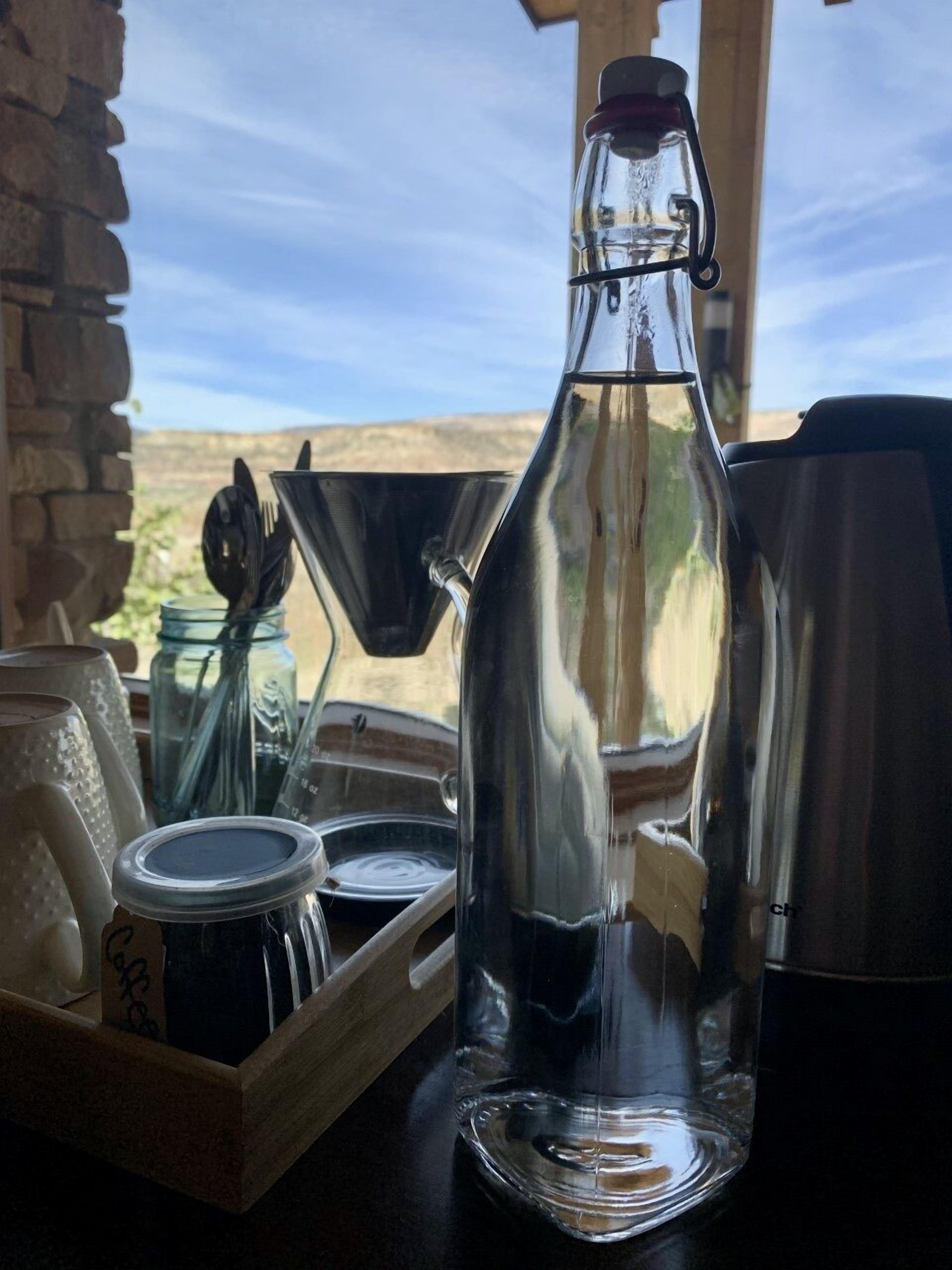 A clear glass bottle with a metal cap, placed on a wooden tray with other glass containers.