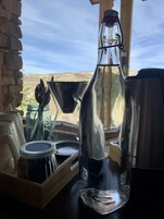 Una botella de vidrio transparente con tapa metálica, sobre una bandeja de madera junto a otros recipientes de vidrio.
