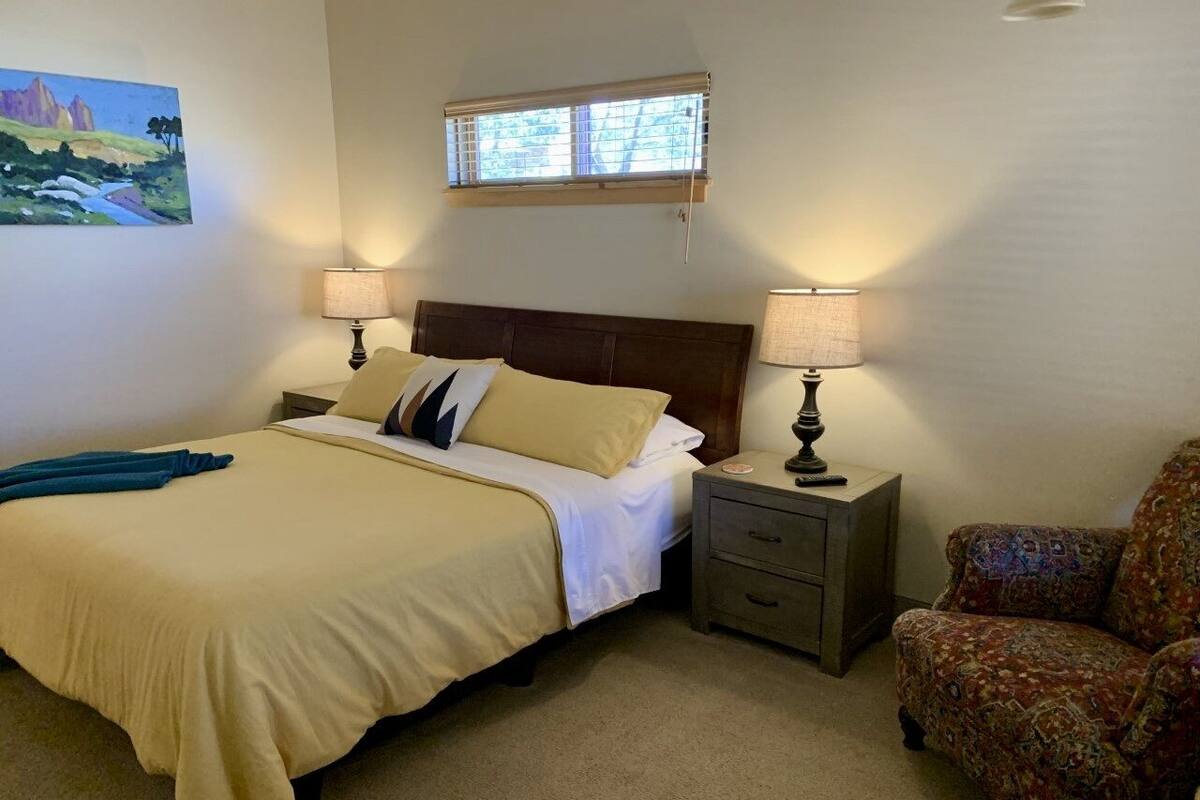 A bedroom with a bed, two bedside tables with lamps, a chair, and a window with blinds.
