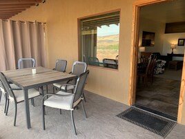 Un patio al aire libre con una mesa y sillas de madera, vistas al paisaje, y una puerta que da a un espacio interior con una lámpara y una silla con estampado de vaca.
