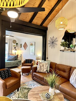 A modern living room with a brown leather sofa, a wooden coffee table, and a patterned rug. There are two armchairs, a floor lamp, and wall art. A bed with a green quilt is visible in the background.