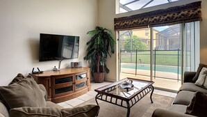 A living room with a flat-screen TV, wooden entertainment unit, glass coffee table, and a large window with a view of a pool and outdoor area. - 4 BR Pool Home/Close to Disney/Universal/Pool/Spa/BBQ/Wifi/Guard Gated Resort (Kissimmee)
