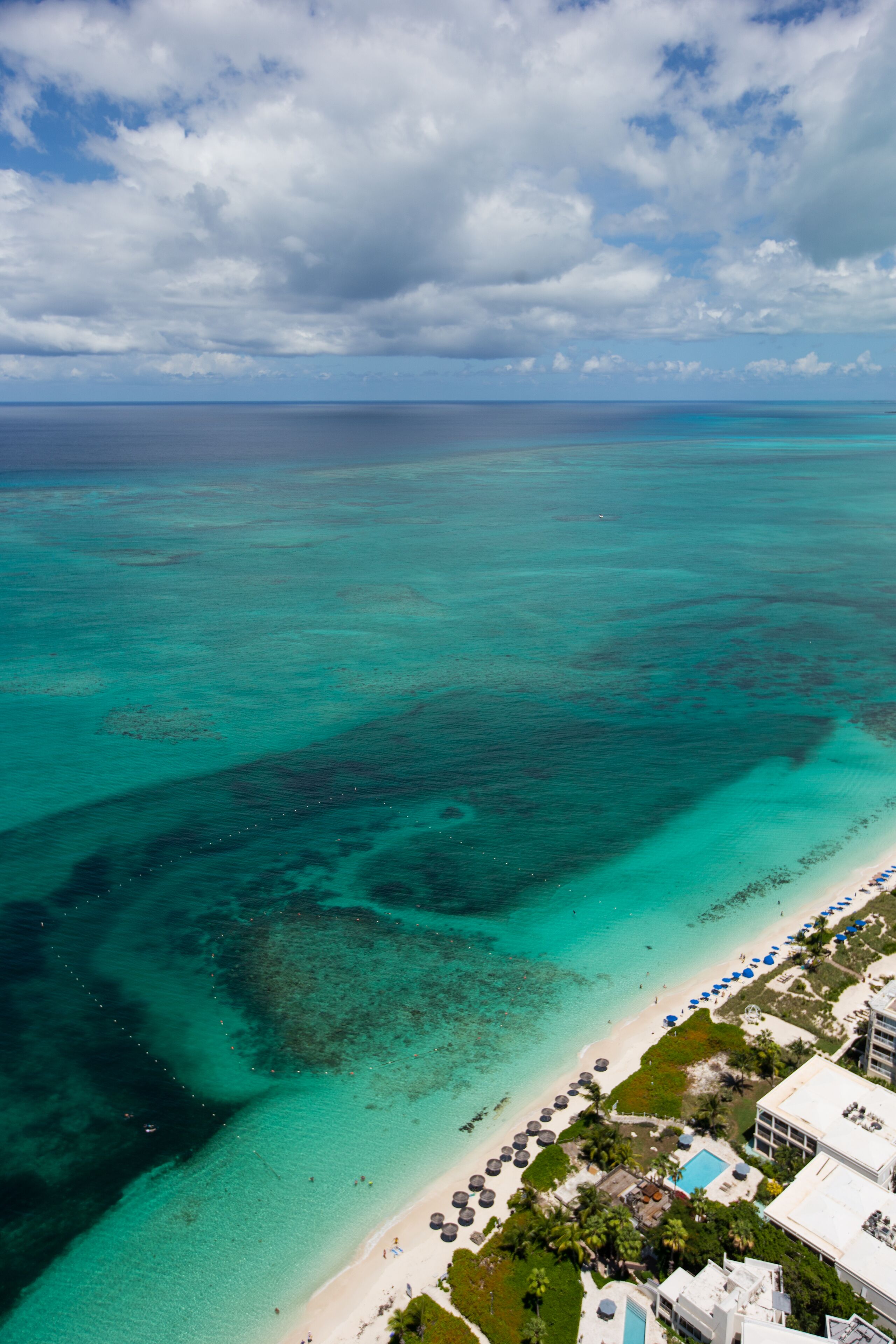 Vista aérea de un complejo turístico en la playa con aguas turquesas cristalinas, una playa de arena y un área de piscina rodeada de palmeras y sombrillas.