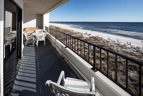 A balcony with white chairs and a table, overlooking a beach with sand and ocean.