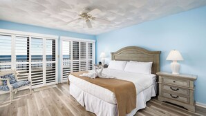A bedroom with a bed, nightstand, lamp, and a view of the ocean through large windows.