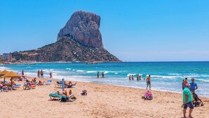 Ein Strand mit klarem, blauem Wasser, Menschen, die sich sonnen, und einer markanten Felsformation im Hintergrund.