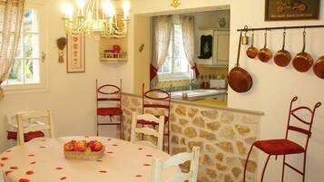 A dining area with a round table covered in a tablecloth, surrounded by white chairs. There is a chandelier above the table, and a stone wall with a kitchen visible through an open door.
