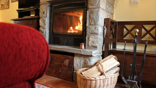 Une cheminée en pierre avec un feu de bois, un banc en bois, un panier en osier rempli de bûches et une lanterne en métal.
