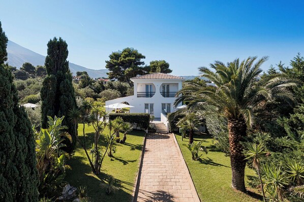 A two-story white house with a tiled roof, surrounded by a well-maintained garden with palm trees and other greenery.