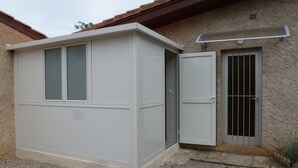 A small, white, single-story building with a gravel driveway, a metal door with a window, and a small vent on the roof.