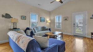 A living room with a sofa, a coffee table, and a ceiling fan. There are two windows with blinds, a door, and a small table with a lamp. - Bonne Mer: Three bedroom, two bathroom house in Oak Island close to shopping and (Oak Island)