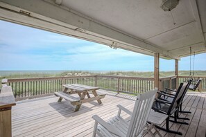 Une terrasse en bois avec une table de pique-nique et des chaises, donnant sur une plage bordée de dunes.