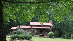 A rustic log cabin with a red roof, surrounded by a well-maintained lawn and a stone circle.