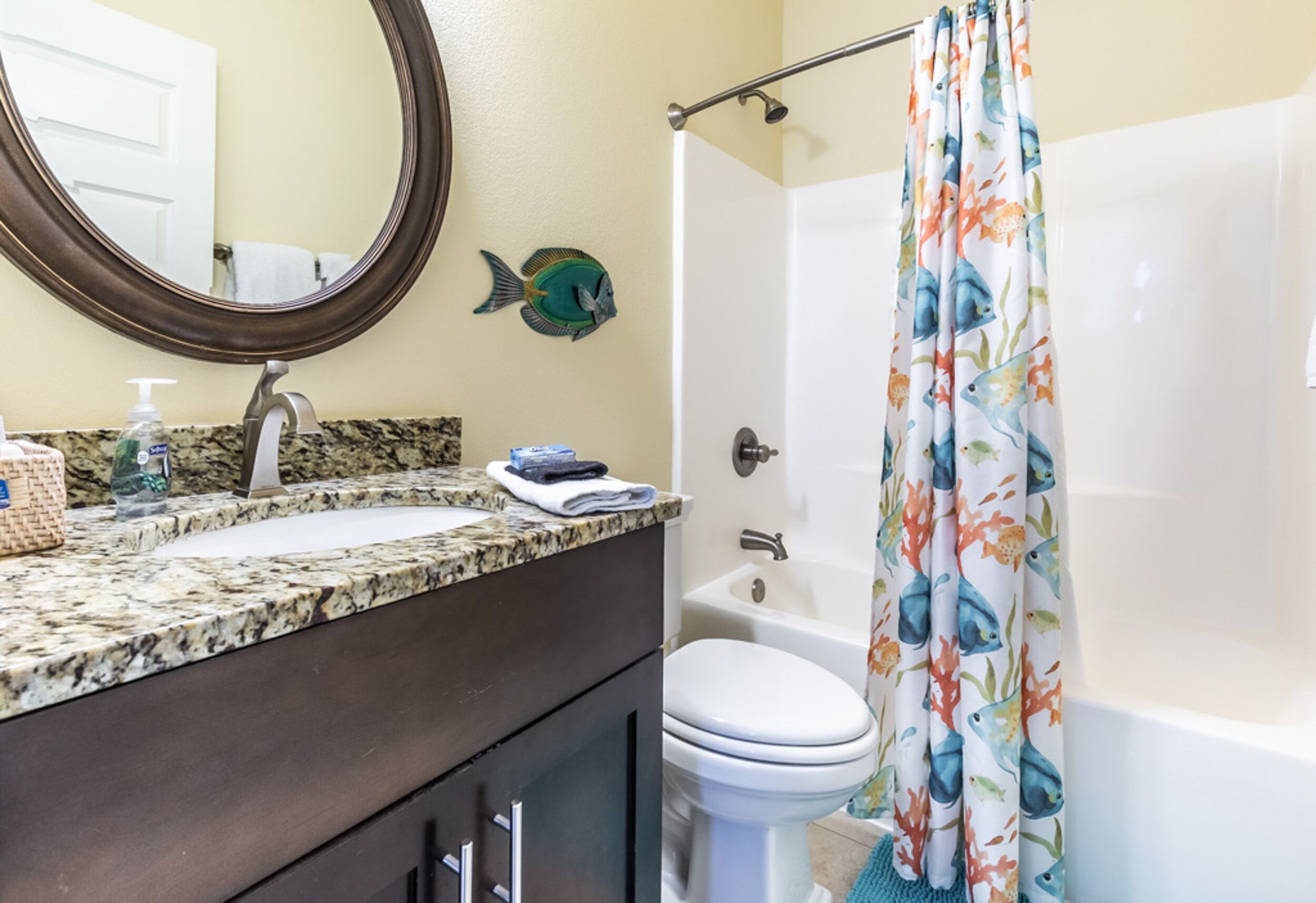 Une salle de bain avec un comptoir en granit, des toilettes blanches, une baignoire et un rideau de douche orné d’un motif de poissons.