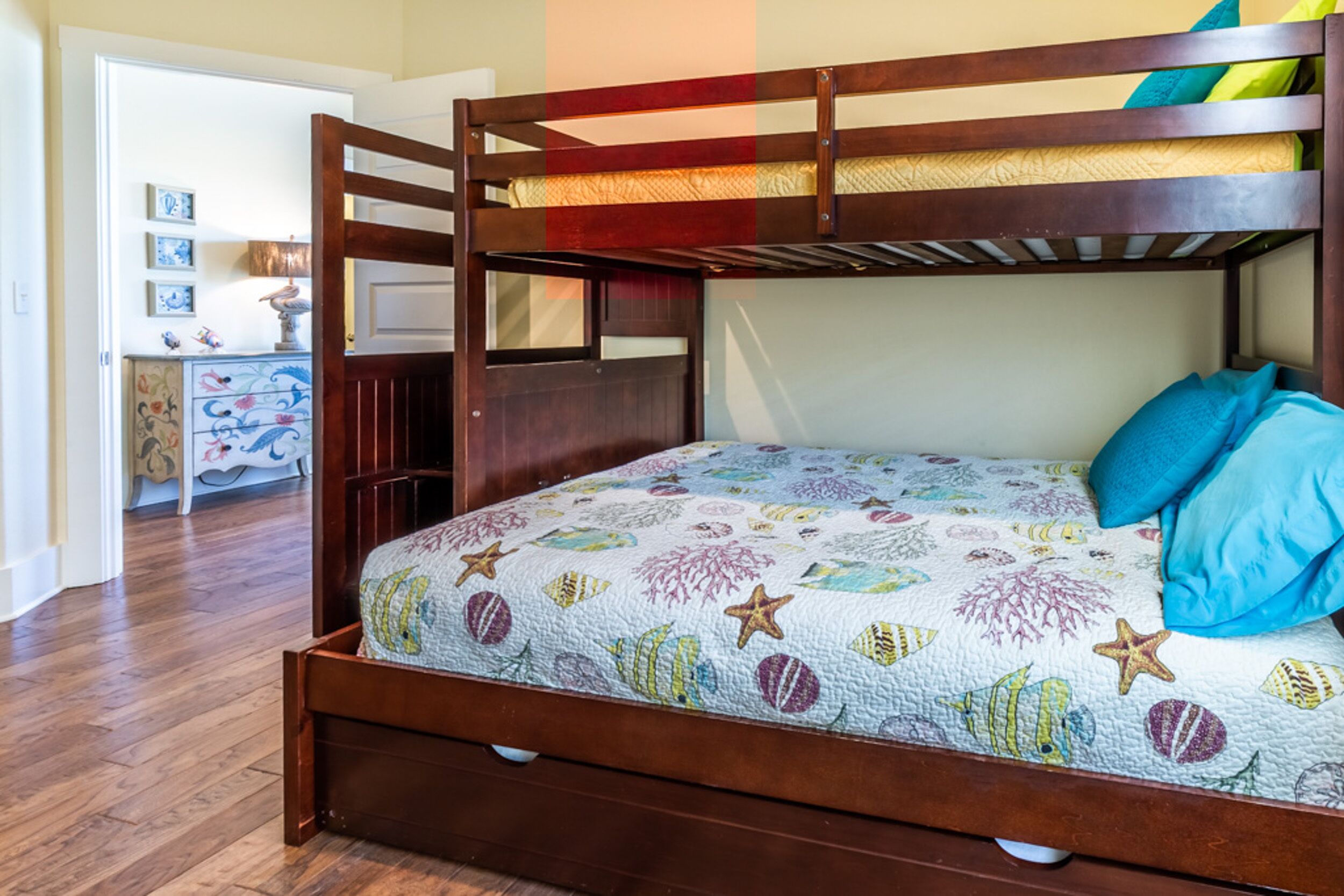 Una cama litera con un edredón colorido, cabecera de madera y un aparador con un mueble de almacenamiento floral al fondo.