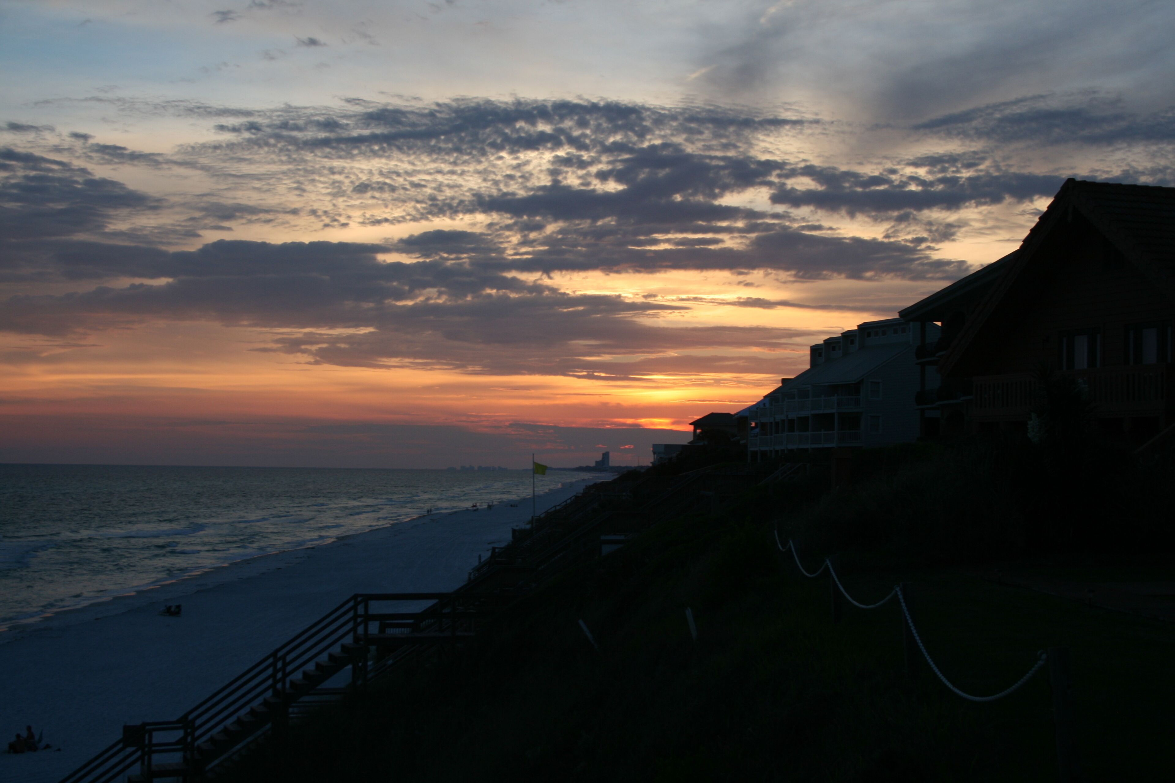A coastal scene with a sunset, a beach, and a building with a balcony overlooking the ocean.