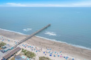 An aerial view of a beach with a long pier extending into the sea, numerous beach umbrellas, and a few buildings along the shore. - Papa's Place: A Stone's Throw to the Beach! (Oak Island)