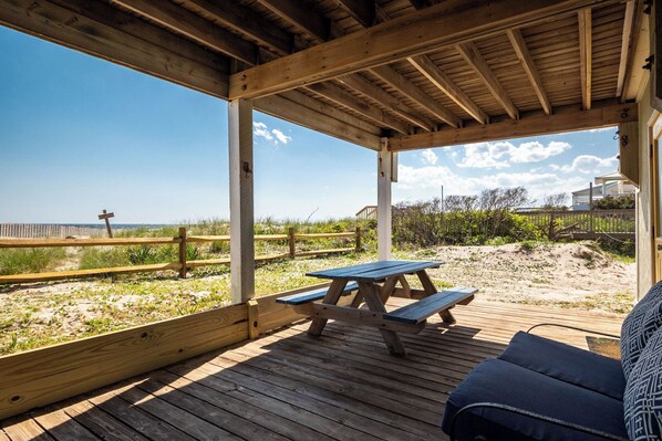 A wooden deck with picnic tables and a bench, overlooking a beach with a wooden fence and a distant beach house. - Beach Barracks: Simple, Oceanfront Home with Large Covered Porch and Dune-top De (Oak Island)