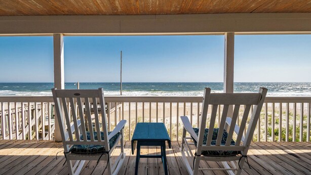 Terraza de madera con dos sillas blancas y una pequeña mesa, con vista a una playa, el océano y un cielo despejado.